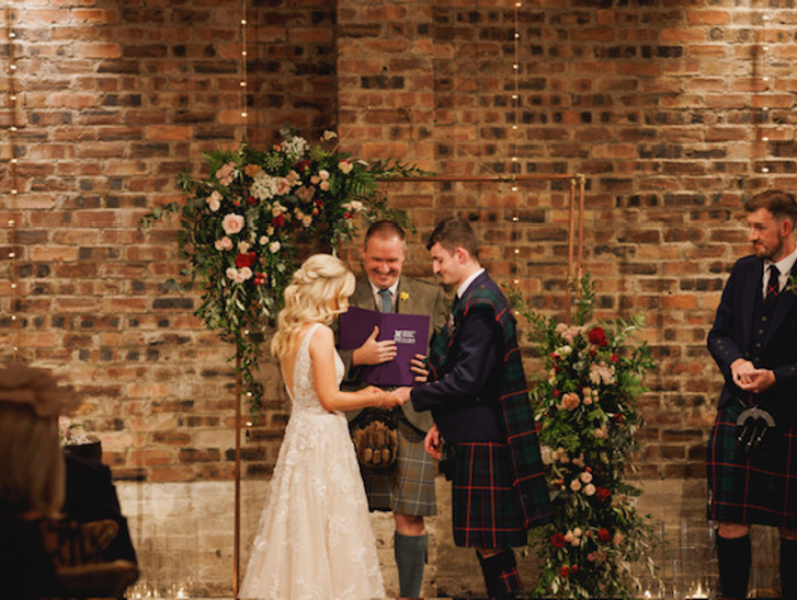 Bride puts grooms ring on his finger whilst celebrant looks on smiling